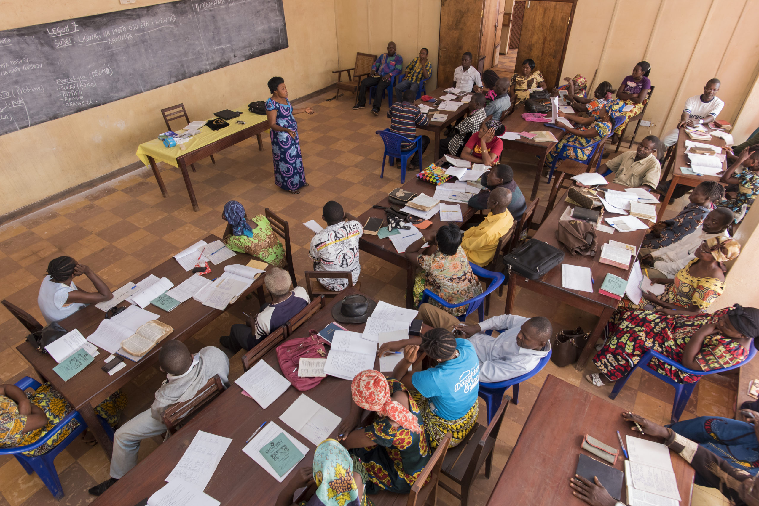 An aerial view of a classroom where people are studying together. A woman speaks at the front.