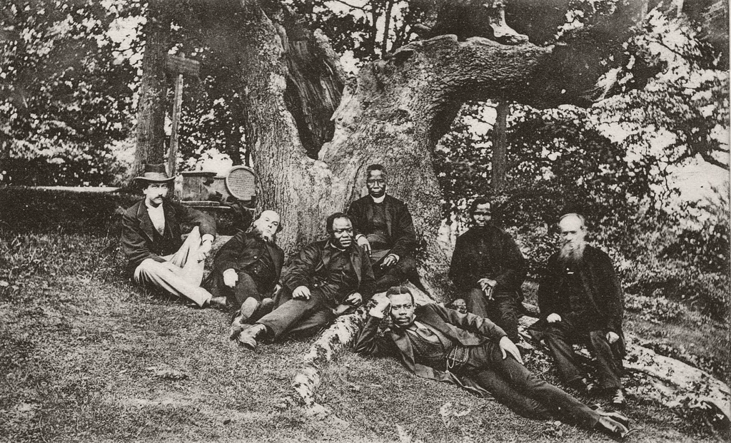 A black and white photo of seven men sitting under a tree together
