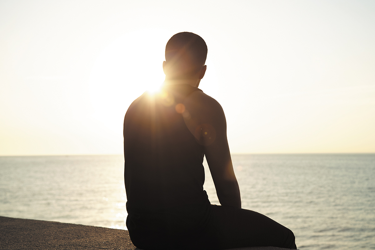 Image of silhouette of pensive young man sitting watching sun setting over sea